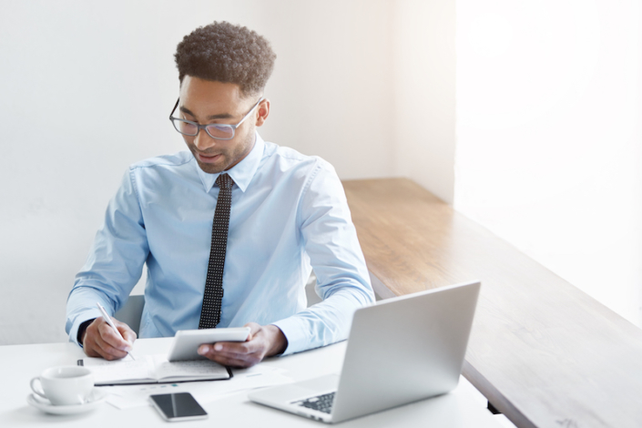 Successful young male dark-skinned accountant with Afro hairstyle and bristle using digital tablet, making notes in copybook, analyzing financial data, sitting at desk surrounded with modern gadgets Accounting and Poor Bookkeeping Tax Liability
